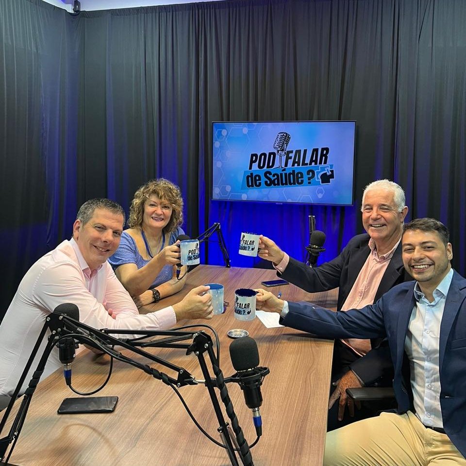 Participantes do PodFalar de Saúde à mesa do estúdio, com canecas do programa e o letreiro do podcast ao fundo.